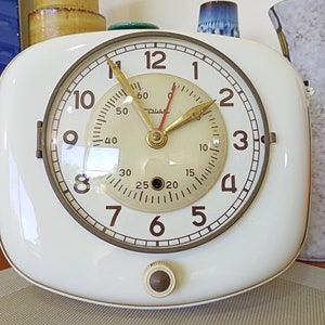 Puede incluir: Un reloj blanco de forma cuadrada con una esfera redonda de color crema. El reloj tiene marcadores de hora con números romanos negros y marcadores de minuto con números arábigos negros. El reloj tiene una aguja de segundos dorada y una aguja de minutos dorada. El reloj tiene un pequeño botón redondo negro en la parte inferior.