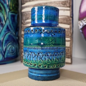 Puede incluir: Un jarrón de cerámica con cuerpo cilíndrico y base acampanada. El jarrón presenta una superficie texturizada con un degradado de color azul y verde. Los elementos decorativos incluyen patrones geométricos y perforaciones. El jarrón mide unos 20 cm de altura.