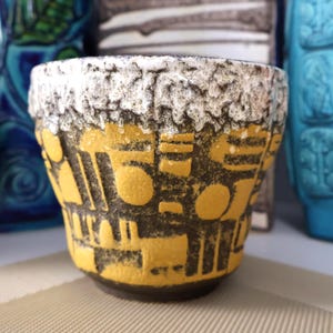 Puede incluir: Un recipiente de cerámica con un borde texturizado blanquecino y un cuerpo amarillo adornado con patrones geométricos negros. El recipiente es probablemente un artículo decorativo o una maceta pequeña. Otros jarrones coloridos son visibles en el fondo.
