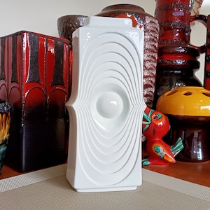 May include: White ceramic vase with a geometric design. The vase features a series of concentric circles that create a ripple effect. The vase is standing on a wooden surface.