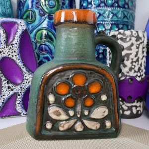 May include: A green ceramic vase with an orange flower design and a handle. The vase has a brown rim and a decorative front panel. Other colourful vases are in the background.
