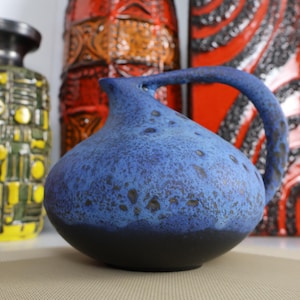 Könnte beinhalten: Ein blauer Keramikkrug mit bauchigem Körper und einem einzelnen Henkel. Die Oberfläche weist eine gesprenkelte Textur auf, mit einem dunkelblauen Boden, der in ein helleres Blau nach oben übergeht. Weitere Keramikgefäße in Gelb, Orange und Rot sind im Hintergrund zu sehen.