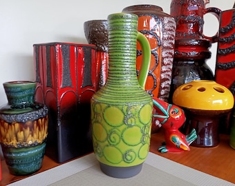 May include: A green and black ceramic vase with a textured surface and a circular pattern. The vase has a handle and a wide mouth.