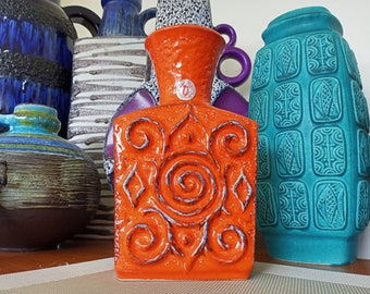 May include: A bright orange ceramic vase with a geometric design. The vase has a square base and a wide opening. The design features a central spiral surrounded by four smaller spirals.