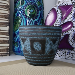 May include: A ceramic planter featuring a light blue and dark brown speckled glaze with a geometric pattern of horizontal bands and triangles. The vessel has a tapered cylindrical shape and a textured finish, positioned in front of other vases.