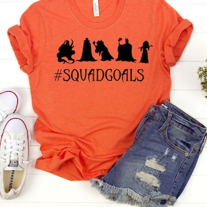 以下が含まれることがあります： 黒いシルエットが5つと「#SQUADGOALS」の文字が入ったオレンジ色のTシャツ。デニムショートパンツと白いスニーカーと合わせています。Tシャツは柔らかく快適な素材で作られています。