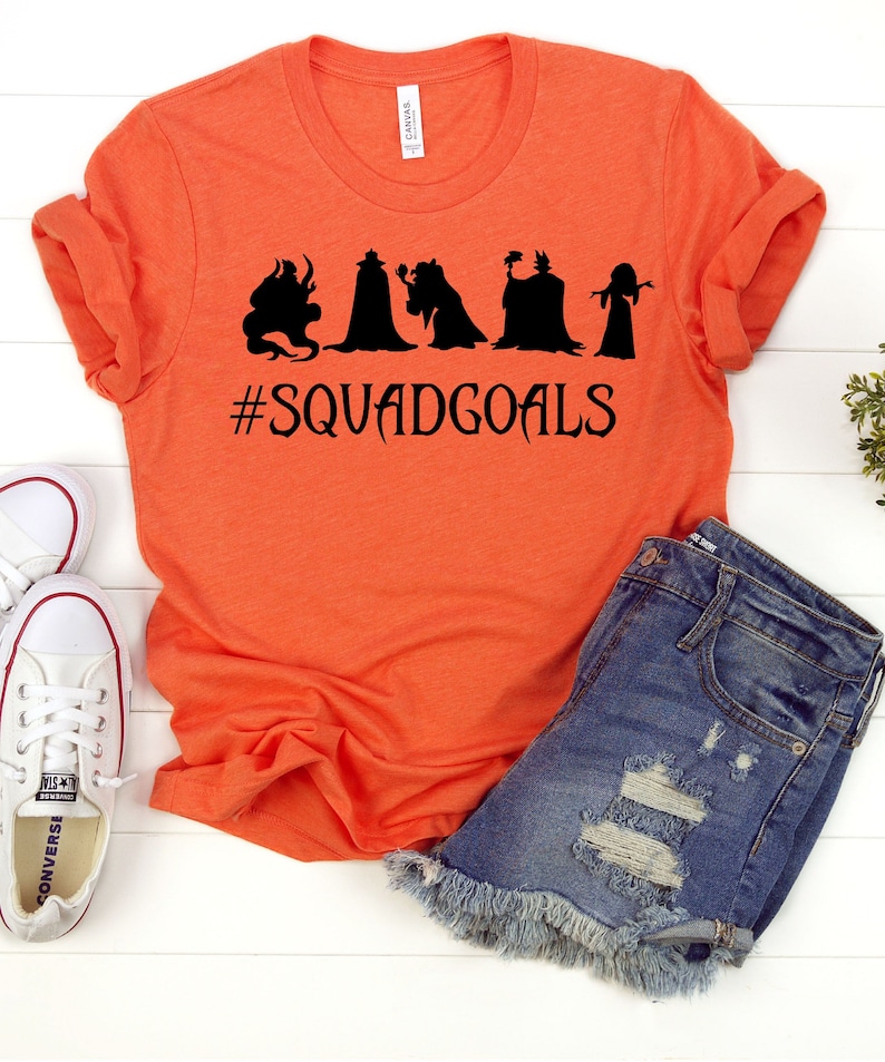 以下が含まれることがあります： 黒いシルエットが5つと「#SQUADGOALS」の文字が入ったオレンジ色のTシャツ。デニムショートパンツと白いスニーカーと合わせています。Tシャツは柔らかく快適な素材で作られています。