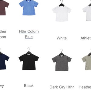 以下が含まれることがあります： ピンク、ブルー、ホワイト、ブラックなど、さまざまな色の半袖Tシャツのセレクション。黒いハンガーに掛けられています。各シャツの下に色の名前がラベル付けされています。これらのシャツはおそらく子供または幼児用です。