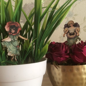 Può includere: Due piante in vaso con bambole di carta decorative. La pianta a sinistra è una pianta verde a foglia in un vaso bianco. La pianta a destra è un bouquet di rose rosse in un vaso di metallo dorato martellato.
