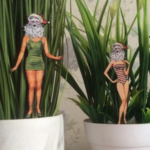 May include: Two paper dolls dressed in swimsuit attire are displayed in potted plants. The dolls have white beards and are wearing Santa hats. The plants are green and leafy.