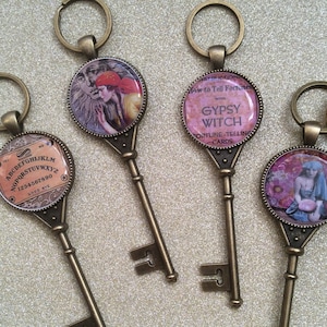 May include: Three antique-style skeleton keys with round glass charms. The charms feature images of a woman in a red hat, a fortune-telling card, and a woman with a crystal ball. The charms are attached to the keys with a metal ring.