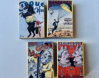 Op de afbeelding: Vier vintage luciferdoosjes met kleurrijke illustraties. De eerste doos heeft een zwart-wit paard met een ruiter, de tweede doos toont een man in een parachute, de derde doos toont een clown met een rode neus en de vierde doos toont twee clowns met rode neuzen.