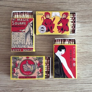 Puede incluir: Cuatro cajas de cerillas vintage con diseños coloridos. La primera caja presenta un diseño rojo y amarillo con el texto "The Magic Square Impregnated" y una cuadrícula de números. La segunda caja tiene un fondo amarillo con dos monos rojos fumando. La tercera caja tiene un fondo rojo con una mujer con un vestido negro y el texto "カフエー旭亭 高崎市歌川町新道". La cuarta caja tiene un fondo amarillo con un pulgar rojo y dos dragones rojos y el texto "MADE IN USA 同公車利國中".
