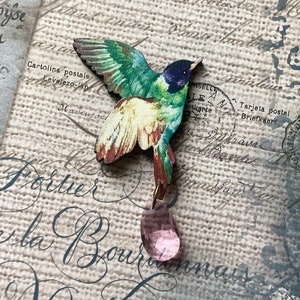 May include: A wooden hummingbird brooch with green, blue, and cream wings, a dark blue head, and a pink crystal teardrop pendant. The brooch is set against a background of vintage postcards and script.