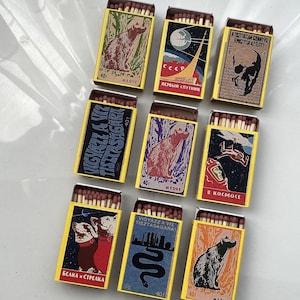 May include: A collection of vintage-style matchboxes with yellow and illustrated designs. The illustrations include a bear, a skull, a rocket, a snake, dogs, and a spaceman. Each box contains wooden matches.