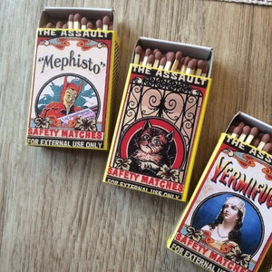 Puede incluir: Tres cajas de cerillas de seguridad con ilustraciones coloridas. La primera caja presenta un diablo rojo con el texto "Mephisto" y "The Assault". La segunda caja presenta un gato negro con el texto "The Assault". La tercera caja presenta una mujer con el texto "Vermifug" y "The Assault".
