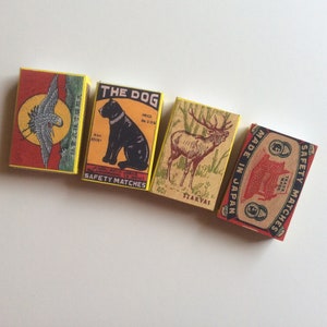 May include: Four vintage matchboxes with colorful illustrations. The first box features a bird with yellow, red, and black colors. The second box features a black dog with the text "The Dog" and "Safety Matches". The third box features a deer with the text "Szarvas" and "Made in Japan". The fourth box features a red and gold design with the text "Safety Matches" and "Trade Mark".
