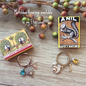 May include: Matchbox keyrings with charms, including a peacock, squirrel, and initial pendants. The image also shows vintage matchboxes with peacock and squirrel illustrations. Text on the image reads "Matchbox Keyrings available in my Etsy Shop."
