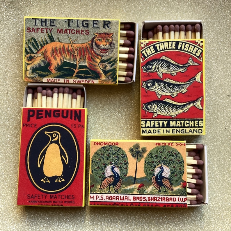 May include: Four vintage matchboxes with different designs. One box features a tiger, another has three fish, a third shows a penguin, and the last depicts peacocks. Each box contains wooden matches with brown tips.