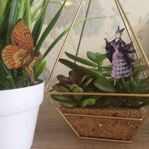 May include: A white potted plant with green leaves and a decorative orange butterfly figurine. A gold geometric terrarium contains succulents and a figurine of a woman in a pink dress and black cape. The terrarium sits on a wooden surface.