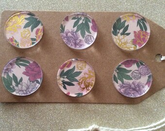 May include: Six round, pink and white floral patterned magnets with gold accents. The magnets are arranged in two rows of three on a brown surface.