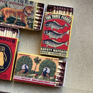 May include: A collection of vintage matchboxes with colorful illustrations. The boxes feature images of a tiger, penguins, fish, and peacocks. The text on the boxes includes "SAFETY MATCHES" and "MADE IN SWEDEN" and "MADE IN ENGLAND".