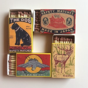May include: Four vintage matchboxes with colorful illustrations. The top box features a red and white illustration of a typewriter with the text "Safety Matches Trade Mark Made in Japan". The box below it features a black and white illustration of a dog with the text "The Dog Price Re 0-0-9 Rho 65057 Je Taraj Match Works Ell Airanrannan Is India Safety Matches". The box to the right features a red and white illustration of a deer with the text "40F Szarvas". The bottom box features a red, white, and yellow illustration of a bird with the text "社會式採寸購同大".
