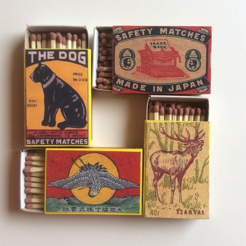 May include: Four vintage matchboxes with colorful illustrations. The top box features a red and white illustration of a typewriter with the text "Safety Matches Trade Mark Made in Japan". The box below it features a black and white illustration of a dog with the text "The Dog Price Re 0-0-9 Rho 65057 Je Taraj Match Works Ell Airanrannan Is India Safety Matches". The box to the right features a red and white illustration of a deer with the text "40F Szarvas". The bottom box features a red, white, and yellow illustration of a bird with the text "社會式採寸購同大".
