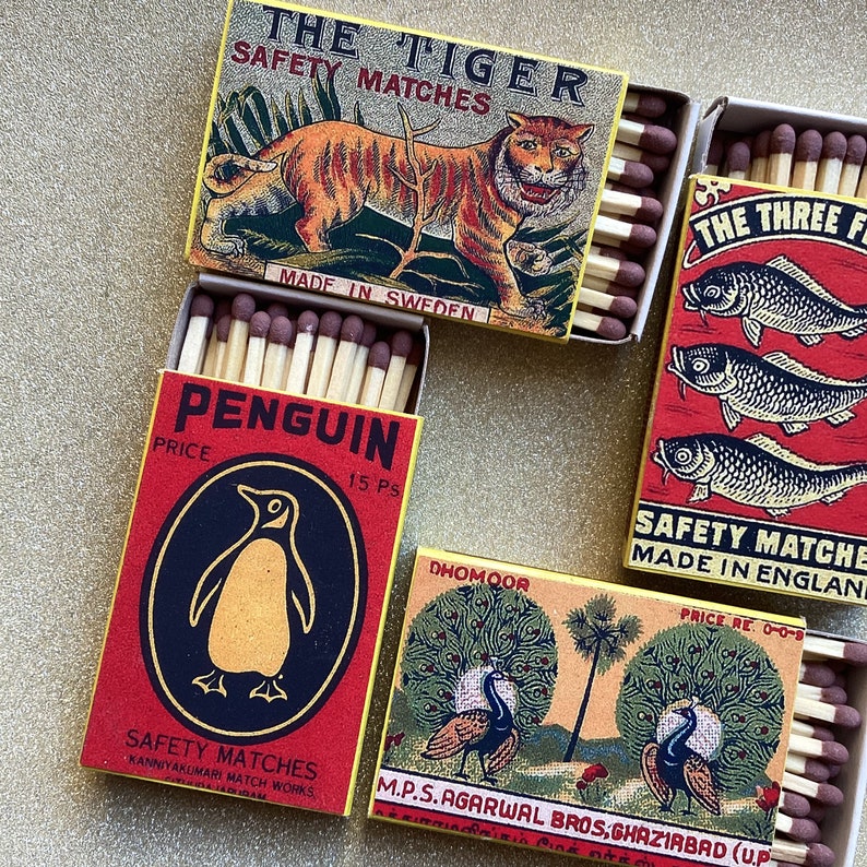 May include: A collection of vintage matchboxes. The boxes feature colorful illustrations of a tiger, fish, penguins, and peacocks. The text on the boxes includes "THE TIGER SAFETY MATCHES", "PENGUIN SAFETY MATCHES", and "THE THREE FISH".