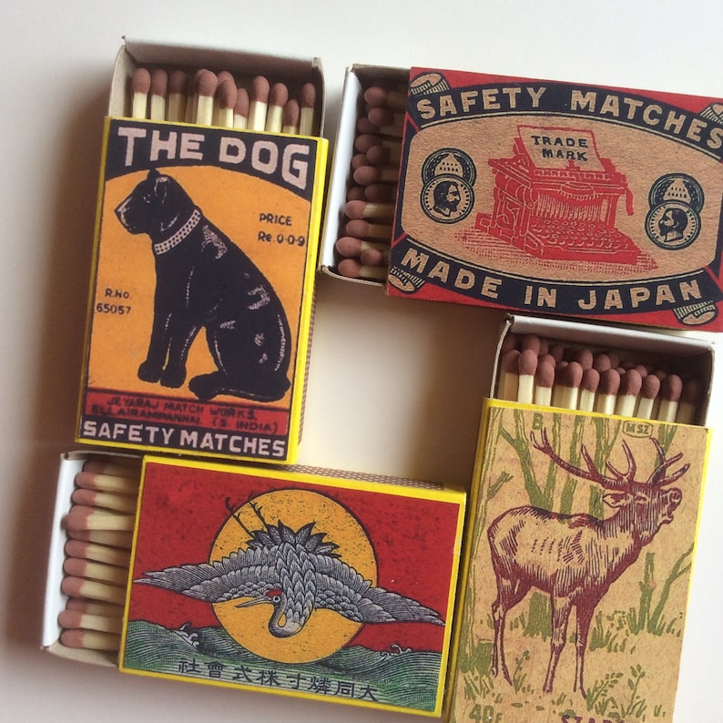 May include: Four vintage matchboxes with different designs. The first box features a black dog with the text "The Dog" and "Safety Matches". The second box has a red and brown design with the text "Safety Matches" and "Made in Japan". The third box has a yellow and red design with a crane and the text "社會式採寸燐周大". The fourth box has a brown and green design with a deer and the text "MSZ".