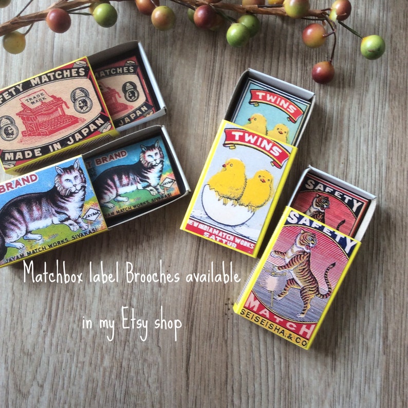 May include: Several vintage matchbox label brooches are displayed, showcasing colorful designs. The brooches feature images of a tiger, cats, and chicks. Text on the boxes includes "Made in Japan" and "Twins". The text "Matchbox label Brooches available in my Etsy shop" is also visible.