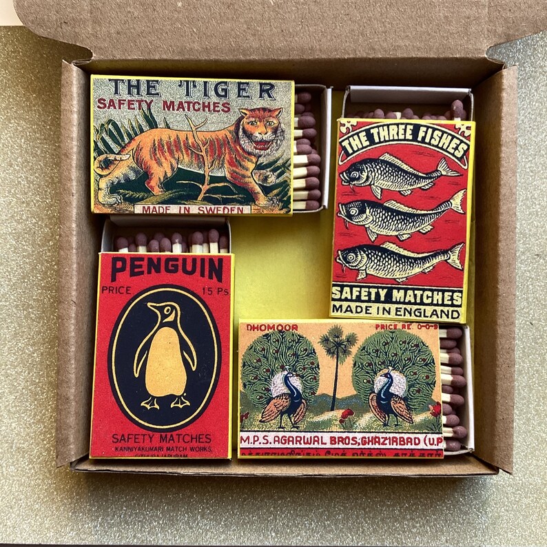 May include: A collection of vintage matchboxes in a cardboard box. The boxes feature colorful illustrations of a tiger, three fish, a penguin, and peacocks. Each box contains wooden matches with red tips.