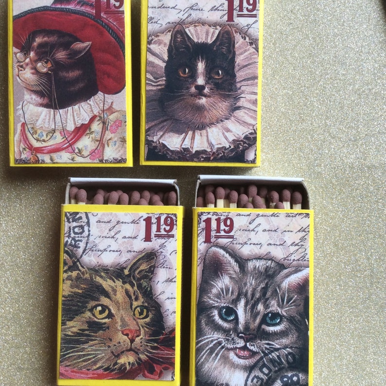 May include: Four matchboxes with yellow and white backgrounds feature vintage illustrations of cats. Each box has the number "119" printed on it. The cats are a black and white cat with a white collar, a brown tabby cat, a gray tabby cat, and a brown and white cat with a red hat.