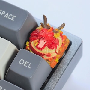 May include: A custom keycap featuring a detailed red dragon design with yellow and orange accents. The keycap is mounted on a gray mechanical keyboard, with the "BACK SPACE", "DEL", and "PAGE UP" keys visible.