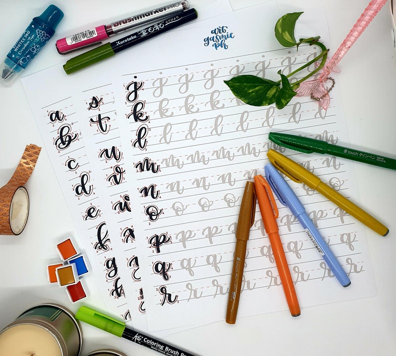 May include: A white sheet of paper with lined sections for practicing calligraphy. The paper has the letters of the alphabet written in cursive at the top of each section. There are four different colored brush pens on the paper, including green, orange, light blue, and brown.