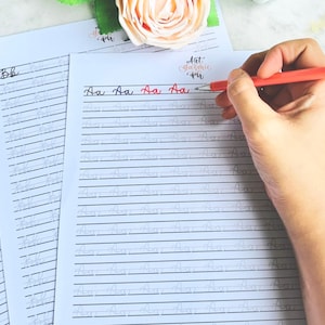 Peut inclure: Une main tenant un stylo rouge écrit sur du papier ligné avec la lettre "Aa" à l'encre rouge. Le papier fait partie d'un ensemble d'entraînement à la calligraphie, avec le texte "Art. Classic. PH" visible. D'autres feuilles avec la lettre "Bb" sont également présentes.