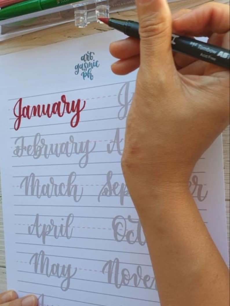 May include: A person's hand holding a black Tombow ABT Dual Brush Pen with a red cap. The pen is being used to write the word "January" in cursive on a white sheet of paper with gray lines. The paper also has the words "February", "March", "April", "May", "September", "October", and "November" written on it.
