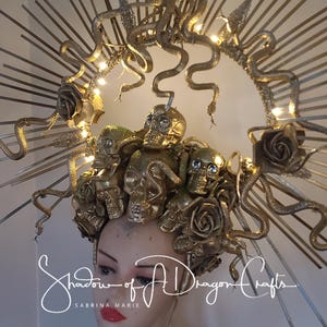 May include: Gold-toned headpiece featuring multiple skull and rose accents, with winding snake details and a halo-like structure. The headpiece is adorned with small lights and the text "Shadow of A Dragon Crafts" is visible.