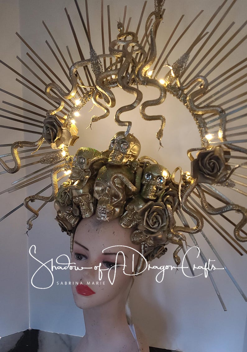 May include: Gold-toned headpiece featuring multiple skull and rose accents, with winding snake details and a halo-like structure. The headpiece is adorned with small lights and the text "Shadow of A Dragon Crafts" is visible.