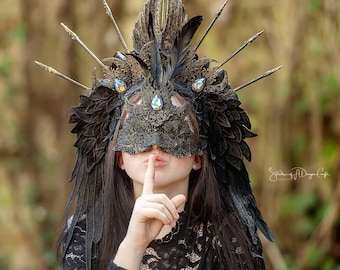 Puede incluir: Una persona con un vestido de encaje negro y una máscara negra y dorada con plumas y joyas. La persona se está llevando el dedo a los labios, como para decir 'shhh'.