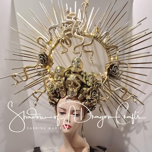 May include: A gold-colored headpiece with a halo design, featuring skulls, roses, and snakes. The headpiece is adorned with radiating spikes and the text "Shadow of a Dragon Crafts" and "Sabrina Marie".