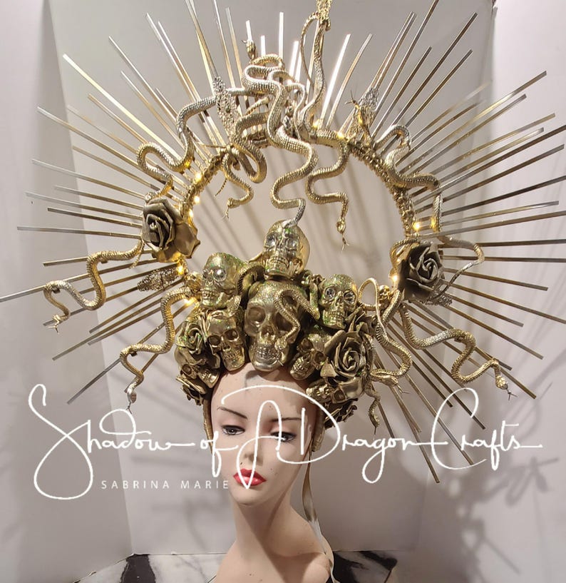 May include: A gold-colored headpiece with a halo design, featuring skulls, roses, and snakes. The headpiece is adorned with radiating spikes and the text "Shadow of a Dragon Crafts" and "Sabrina Marie".