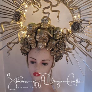 May include: A golden headpiece featuring skulls, roses, and snakes. The crown is adorned with decorative elements and lights, creating a dramatic effect. The headpiece is displayed on a mannequin head with red lipstick. The text "Shadow of A Dragon Crafts" is visible.