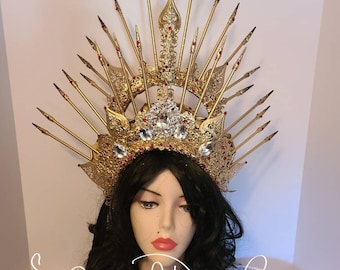 May include: A gold-colored crown with intricate detailing, adorned with red and clear gemstones. The crown features multiple upward-pointing spikes and is displayed on a mannequin head with black hair. The text "Shadow of A Dragon Crafts" is visible.