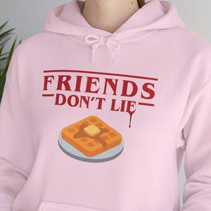 以下が含まれることがあります： フロントポケットとドローストリングフードが付いた、薄いピンク色のパーカー。「FRIENDS DON'T LIE」の赤い文字と、バターを添えたワッフルのイラストが前面に描かれています。