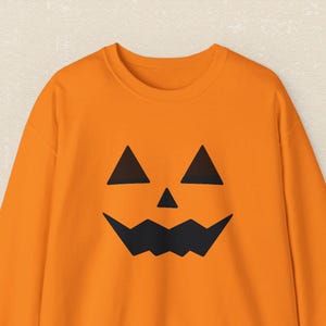 Może przedstawiać: Pomarańczowa bluza z czarnym wzorem twarzy dyni. Bluza o tematyce Halloween ma klasyczną twarz dyni z trójkątnymi oczami i poszarpanymi ustami. Odświętny i wygodny element garderoby na straszny sezon.