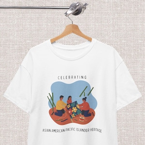 Peut inclure: T-shirt blanc avec un graphisme célébrant le patrimoine des Américains d'origine asiatique et des insulaires du Pacifique. Le design présente trois personnages, des fleurs et le texte "CELEBRATING ASIAN AMERICAN PACIFIC ISLANDER HERITAGE" en noir.