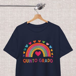 May include: A navy blue short-sleeved t-shirt featuring a colourful graphic of a rainbow with a pink heart in the centre, surrounded by small hearts. The text QUINTO GRADO is printed in multicoloured letters below the rainbow design.