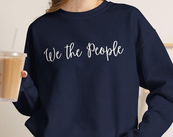 May include: A navy blue long-sleeve crewneck sweatshirt featuring the white text "We the People" in a cursive script font across the chest. The sweatshirt is paired with gray denim shorts, creating a casual and simple outfit combination.