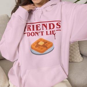 May include: A light pink hooded sweatshirt featuring a graphic print with the red text "FRIENDS DON'T LIE" above an illustration of a waffle with a pat of butter on a plate. The hoodie includes a front kangaroo pocket and matching drawstrings.